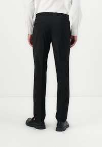 Pantalon noir avec une coupe ajustée, en tissu lisse, avec une taille à passants de ceinture et deux poches arrière. Porté avec des chaussures noires.