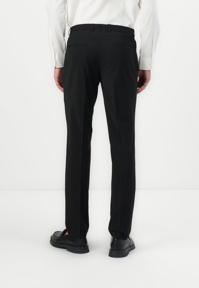 Pantalon noir avec une coupe ajustée, en tissu lisse, avec une taille à passants de ceinture et deux poches arrière. Porté avec des chaussures noires.