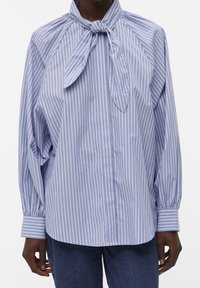 Blusa a righe blu e bianche con collo a fiocco, maniche lunghe e chiusura con bottoni. Il tessuto ha una texture liscia con una vestibilità comoda.