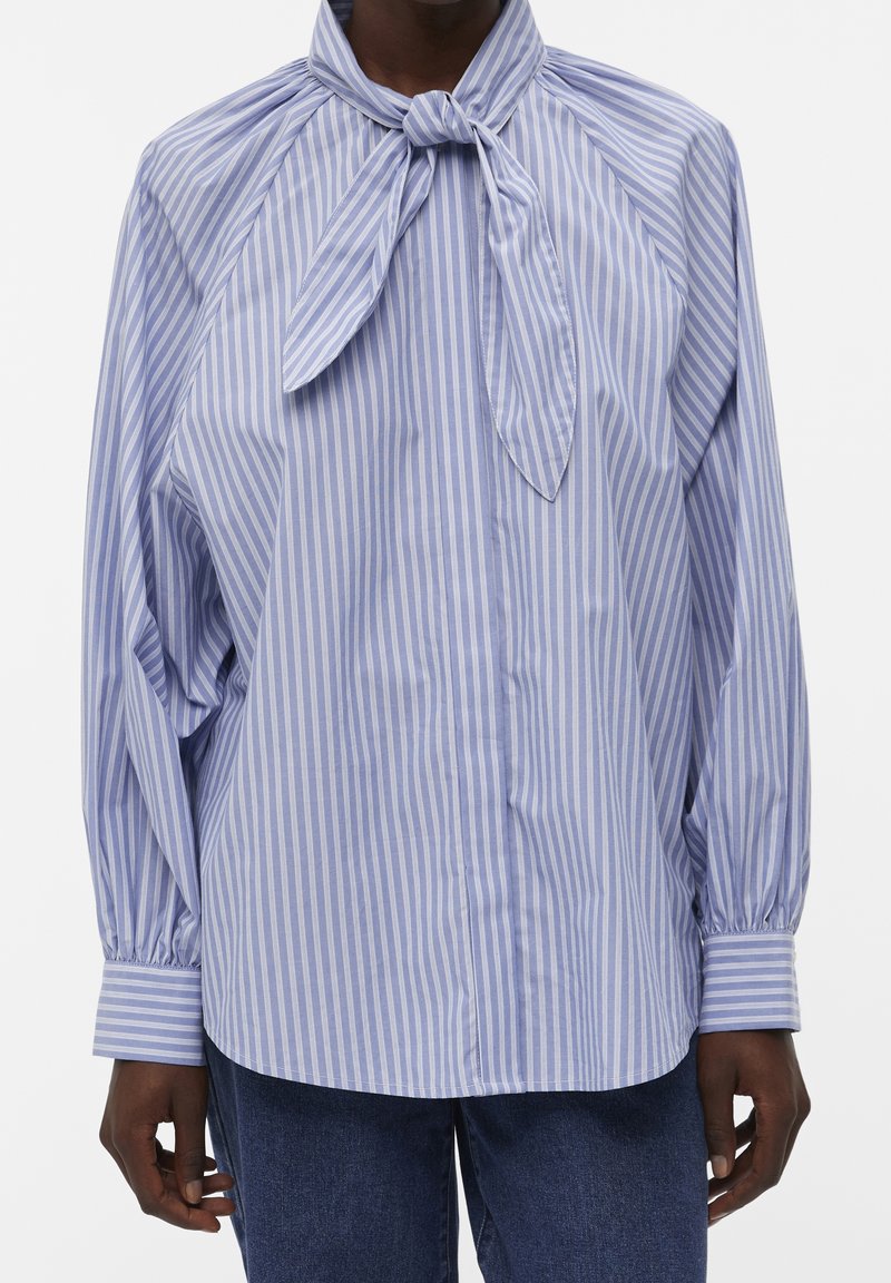 Blusa a righe blu e bianche con collo a fiocco, maniche lunghe e chiusura con bottoni. Il tessuto ha una texture liscia con una vestibilità comoda.