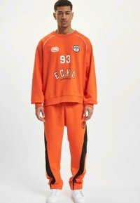 Orange sweatshirt and matching sweatpants with black side stripes. Features white text, including "ECKÖ" and number "93." Made of soft fabric.