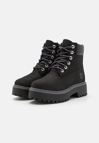 Sorte anklestøvler i ruskind med en tyk, robust sål, metaløjer og grå snøringer, med logo detaljer på siden.