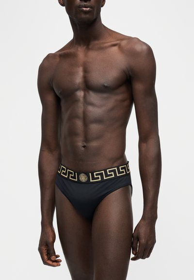 Homme torse nu portant un slip noir à ceinture motif Greca doré et emblème Méduse, debout devant un fond uni clair.