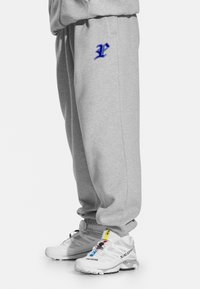 Szare spodnie dresowe z haftowanym niebieskim logo na lewej udzie. Noszone z białymi butami sportowymi z wielokolorowymi akcentami i teksturowaną podeszwą.