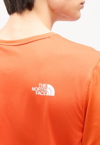 Άτομο που φοράει πορτοκαλί πουκάμισο με υφή, με το λογότυπο The North Face τυπωμένο με άσπρο χρώμα στο πάνω μέρος της πλάτης, στραμμένο μακριά από την κάμερα.