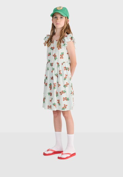 Dievča v zelenej čiapke, bielych šatách s oranžovými vzormi čerešní, bielych ponožkách a červených žabkách stojí so založenými rukami na jednoduchom pozadí.