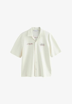 Krémová košeľa s krátkym rukávom na gombíky s golierom a červeným potlačeným textom na ľavej a pravej strane hrudníka.