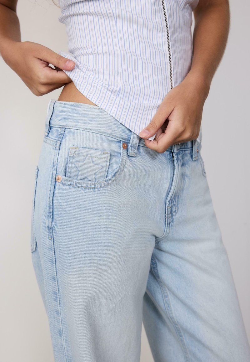 Personne ajustant un jean bleu clair avec un motif d’étoile sur la poche, portant un haut sans manches blanc et bleu rayé.