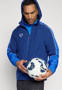 Γαλάζια ελαφριά μπουφάν με κουκούλα, λευκές λεπτομέρειες και ελαστικούς καρπούς. Το μοντέλο κρατάει μια μπάλα ποδοσφαίρου σε λευκό και γαλάζιο, φορώντας μια γαλάζια μπλούζα από κάτω.