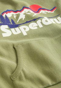 Sudadera de color verde oliva con un prominente logo en blanco de "Superdry" y un gráfico de paisaje alpino con un sol rojo y contornos azules.