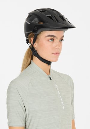 Sort cykelhjelm med ventilationshuller, justerbar rem og en mat overflade, båret med en lys grøn struktureret skjorte med en lynlås foran.
