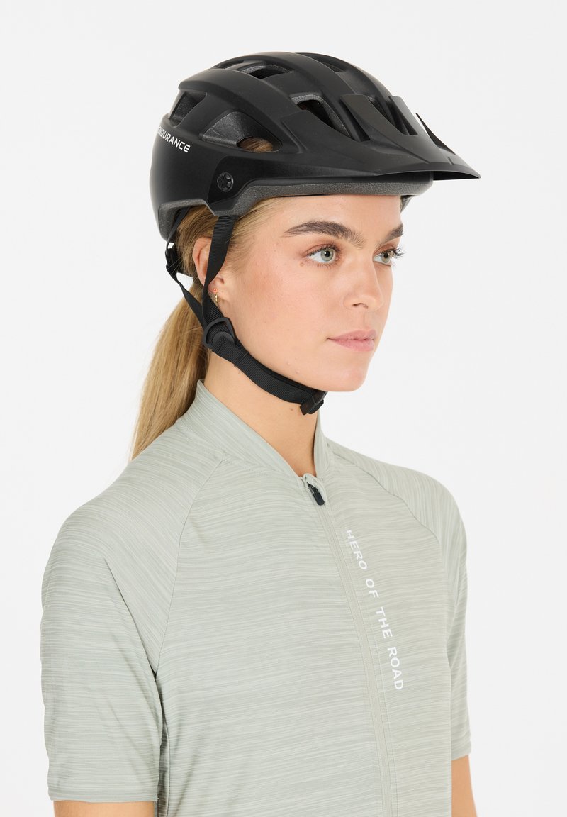 Schwarzer Fahrradhelm mit Belüftungsöffnungen, verstellbarem Riemen und matter Oberfläche, getragen mit einem hellgrünen, strukturierten Shirt mit einem Frontreißverschluss.