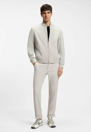 Junger Mann trägt eine hellbeige Bomberjacke und Hose, darunter ein schwarzes Hemd, steht mit den Händen in den Taschen, weiße und schwarze Sneaker.