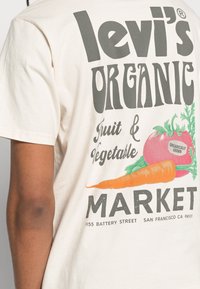 Krémové tričko s velkým grafickým designem. Text zní "levi's ORGANIC," s obrázky mrkve a rajčete, a detaily z trhu.