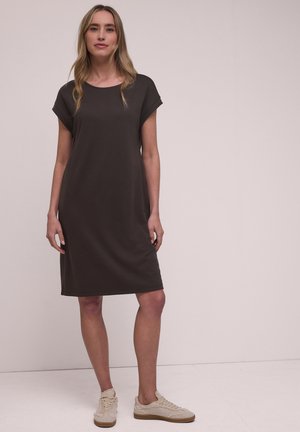 Kvinde stående iført en løs, mørkebrun kjole til knæene og beige sneakers mod en ensartet lys baggrund.