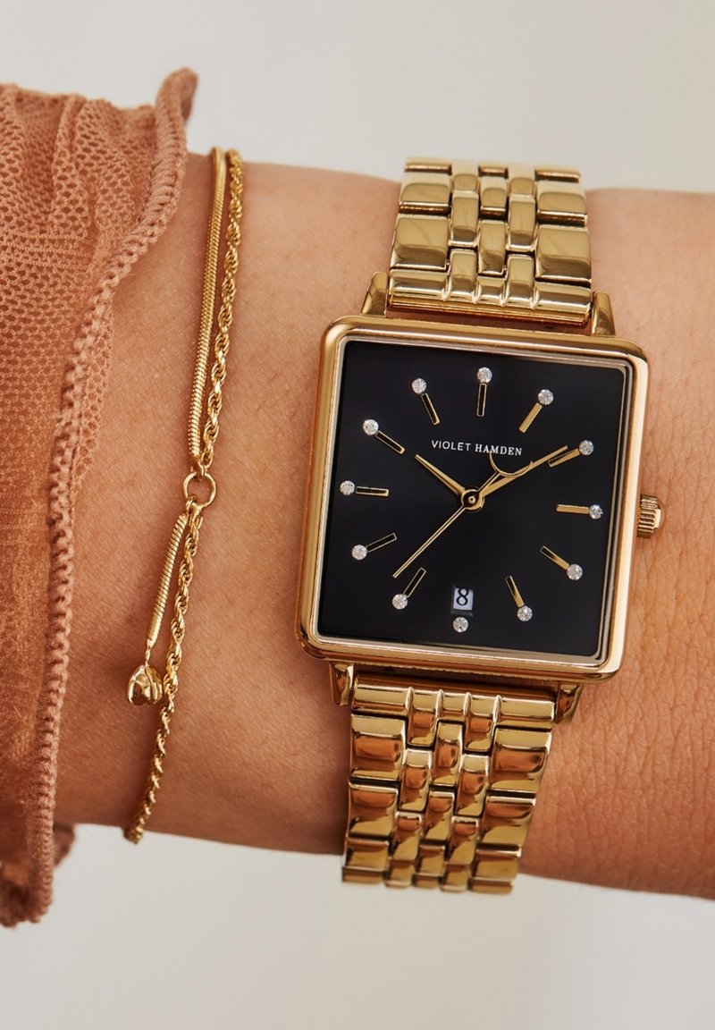 Gouden rechthoekige horloge met een zwarte wijzerplaat, diamantachtige uurmarkeringen, en een gouden metalen armband. Vergezeld van een dunne gouden kettingarmband.
