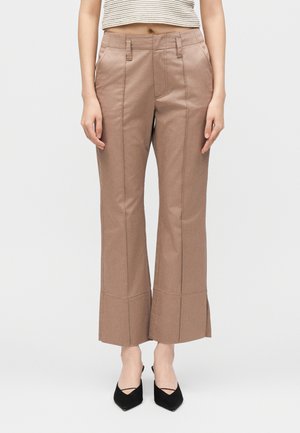 Γυναίκα που φοράει καφέ παντελόνες με ψηλή μέση και έντονες ραφές μπροστά, συνδυασμένες με μαύρες μυτερές γόβες και κοντό ριγέ τοπ.