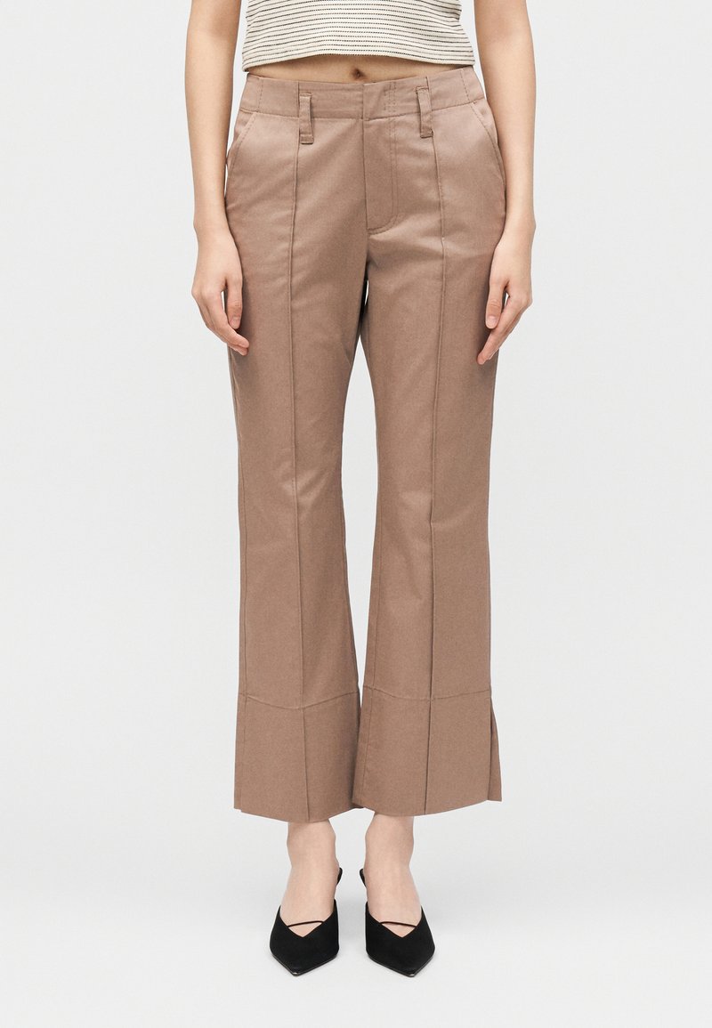 Γυναίκα που φοράει καφέ παντελόνες με ψηλή μέση και έντονες ραφές μπροστά, συνδυασμένες με μαύρες μυτερές γόβες και κοντό ριγέ τοπ.