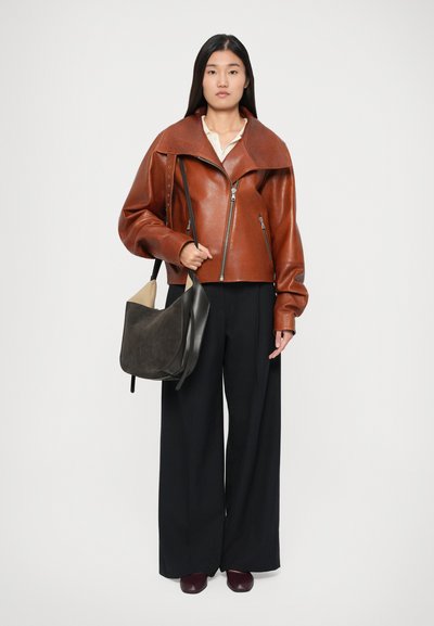 Giacca di pelle marrone con ampi revers, chiusura con zip e maniche lunghe; abbinata a pantaloni neri a gamba larga e una borsa a spalla grigio scuro.