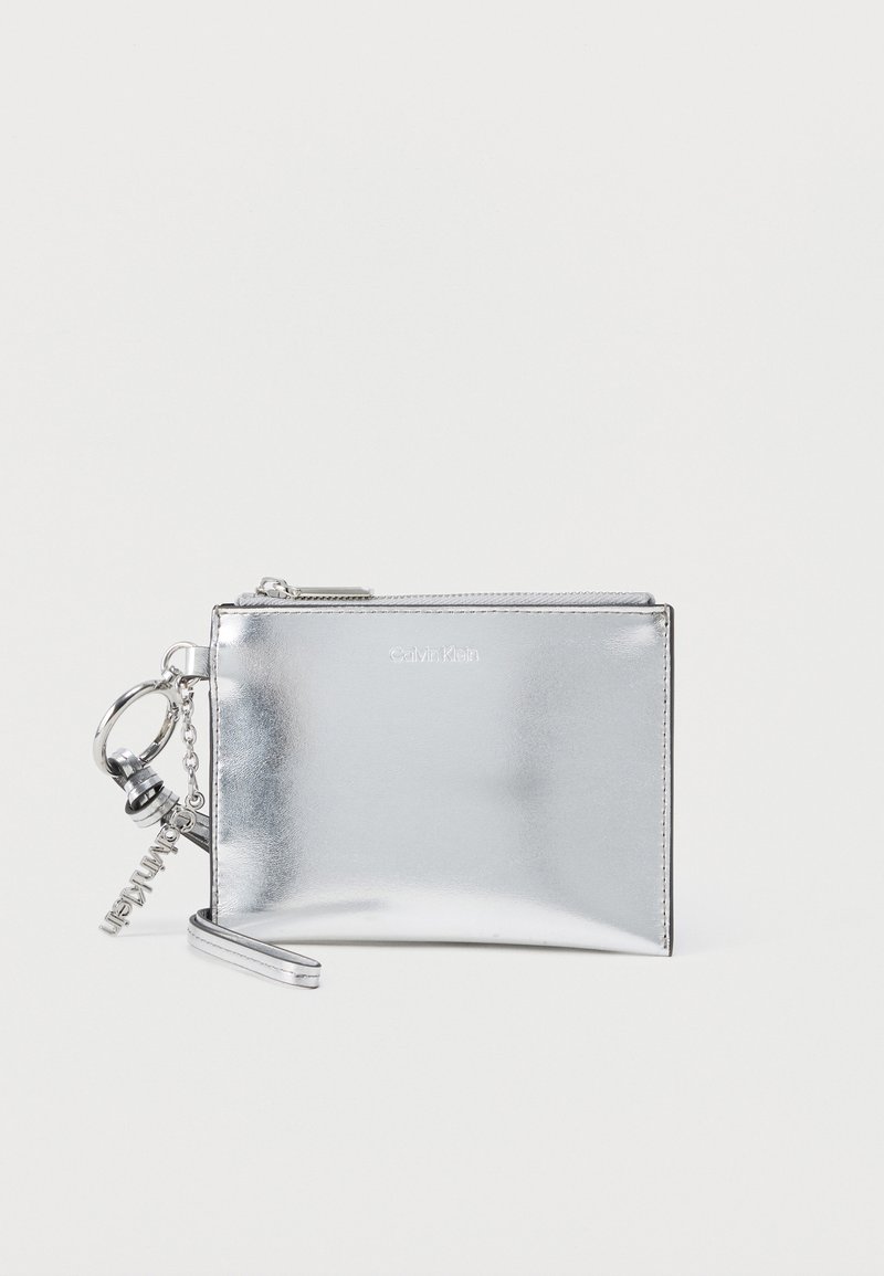 Ασημί μεταλλικό πορτοφόλι χειρός Calvin Klein με φερμουάρ και διακριτικό μπρελόκ με λογότυπο σε απλό φόντο.