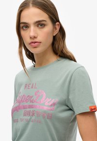 Camiseta de algodón verde claro con mangas cortas, que presenta el texto en gliter rosa "REAL SuperDry" y una pequeña etiqueta de logo naranja en la manga.