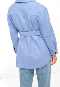 Světle modrá košilová bunda z vlněné směsi s páskem v pase, knoflíkovými manžetami a klopami vzadu, kombinovaná se světlou denimovou kalhoty.