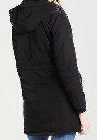 Černá prošívaná bunda s kapucí, dlouhými rukávy a staženým pasem. Hladká textura a lem, který sahá do poloviny stehna.