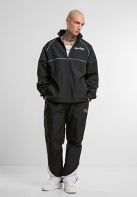 Veste coupe-vent noire avec zip, dotée d'un logo blanc et d'accents bleu clair. Associée à un pantalon assorti et des baskets blanches. Texture de tissu mate.