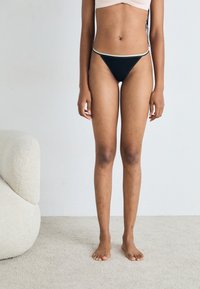 Chiloți negri de bikini cu un brâu subțire, accentuat cu detalii aurii. Design cu acoperire minimă și textură netedă. Modelul stă desculț.