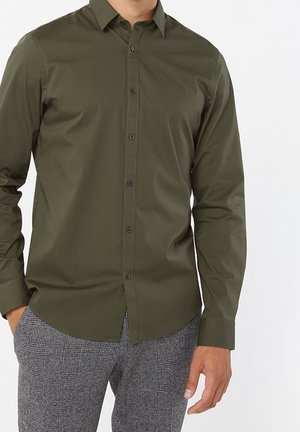 Mann trägt olivegrünes Hemd mit Knöpfen und langen Ärmeln sowie graue karierte Hose, steht mit teilweise in den Taschen gesteckten Händen.