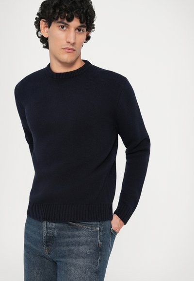 Uomo che indossa un maglione a girocollo blu scuro e jeans blu, in piedi con una mano in tasca su uno sfondo bianco semplice.