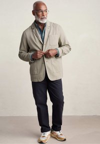 Blazer in lino beige con una tasca, abbinato a una camicia a quadri blu, pantaloni blu navy e sneakers multicolori.