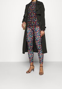 Person mit passendem buntem, blumigen Langarmoberteil und Leggings, schwarzem Trenchcoat, schwarzen Knöchelriemchen-Pumps und hält eine kleine goldene Handtasche.