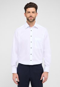 Weißes Langarmshirt mit einem Button-Down-Kragen, ausgestattet mit einer einzelnen Brusttasche und dunklen Knöpfen auf einem glatten Stoff.