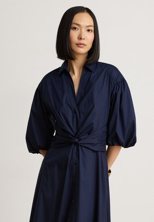 Jonge vrouw met stijl zwart haar die een marineblauwe jurk draagt met pofmouwen en een gedraaide knoopdetails in de taille.