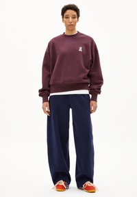 Weinrotes Sweatshirt mit einem kleinen weißen Schleifenlogo, kombiniert mit marineblauen weiten Hosen und mehrfarbigen Sneakers mit orangefarbenen Akzenten und Schnürsenkeln.