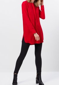 Tunique rouge à manches longues avec un col rond et un plastron de boutons, associée à des leggings texturés noirs et des bottines.