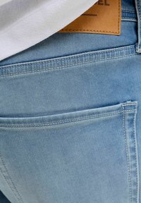 Κοντινό πλάνο σε τσέπη ανοιχτό μπλε τζιν με ορατή λευκή ραφή και μερική καφέ δερμάτινη ετικέτα κάτω από άσπρο ύφασμα.