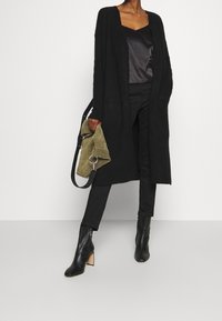 Long black cardigan made of textured fabric, paired with a satin black camisole, black trousers, and black heeled boots. Holding a green bag.