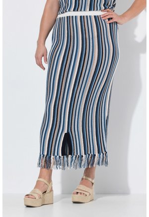 Žena nosící modrou, bílou a béžovou proužkovanou pletenou sukni s třásněmi na lemu a rozparkem vpředu, doplněnou béžovými tkanými platformními sandály.