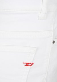 Tessuto denim bianco con una texture liscia. Presenta un logo ricamato rosso sulla tasca posteriore, cuciture bianche e dettagli di cucitura aggiuntivi.
