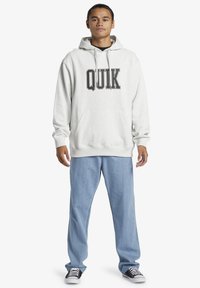 Lys grå sweatshirt med "QUIK" i fed sort skrift, parret med løse lyseblå jeans og sorte sneakers. Har en frontlomme.