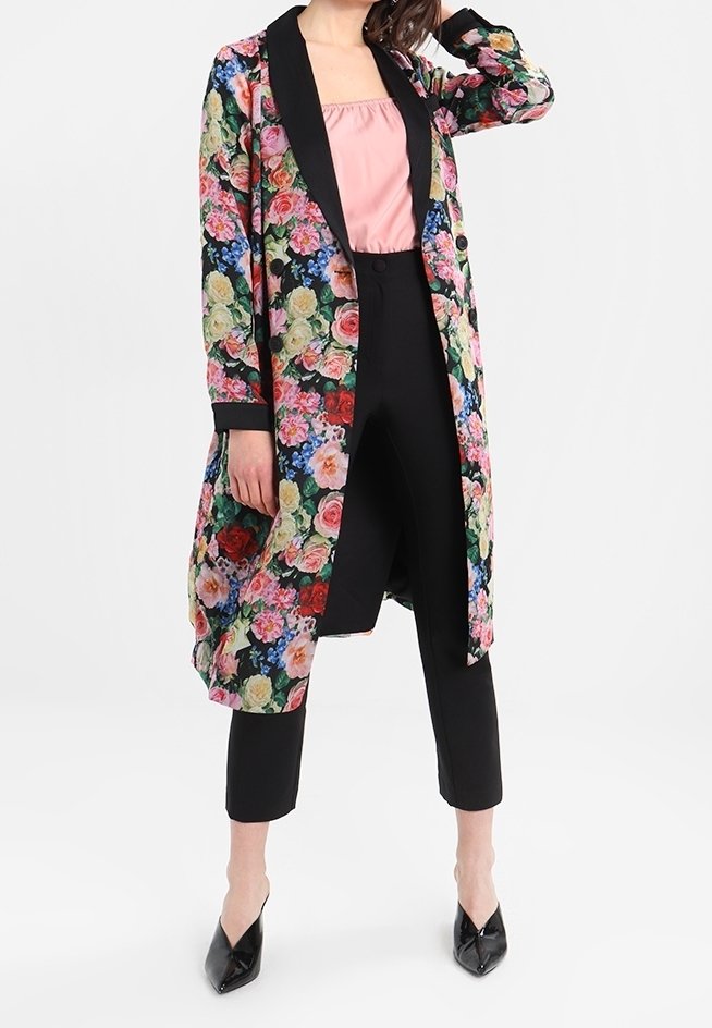Manteau long à motifs floraux avec revers noirs, mettant en valeur des roses vives et des fleurs bleues. Associé à un pantalon noir et un haut rose clair.