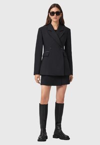 Zwarte dubbelzijdige blazer met een op maat gemaakte pasvorm, gecombineerd met een bijpassende minirok. Accessoires zijn onder andere zwarte zonnebrillen en kniehoge laarzen.