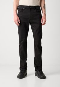 Zwarte katoenbroek met een slim fit, vijf zakken, sluiting met een knoop aan de voorkant en subtiele branding op de voorzak. Gekleed met zwarte sneakers.