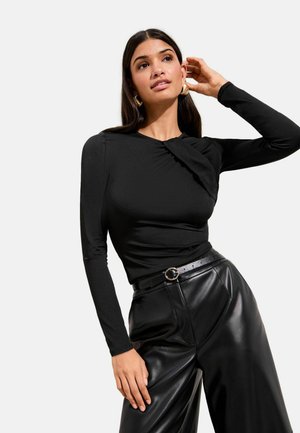 Top negru cu mâneci lungi, cu detaliu adunat pe un umăr, asortat cu pantaloni negri din piele cu talie înaltă și o curea neagră subțire cu o cataramă circulară.