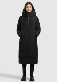 Lang sort pufferjakke med høj krave, chevron-quiltet mønster, lynlåse og sidelommer. Båret med sorte leggings og ankelstøvler.