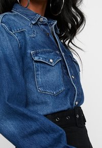 Chemise en denim bleu foncé avec un col rabattu, deux poches poitrine, fermeture à boutons-pression et manches longues. Bas taille haute noir avec ceinture visible.