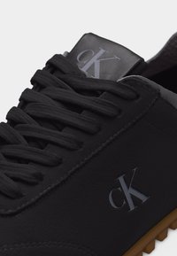 Baskets noires avec lacets noirs, logo "cK" gris sur le côté et la languette, et une semelle crantée marron.