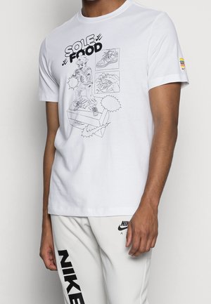Mężczyzna w białej koszulce Nike z rysunkiem w stylu kreskówkowym przedstawiającym osobę na pudełku na buty, szkicem sneakersa i tekstem „SOLE FOOD” napisanym pogrubioną czcionką.